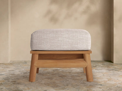 Maxime Outdoor Ottoman with a light gray textured cushion on a natural teak wood base, set on a stone patio with a light beige wall.