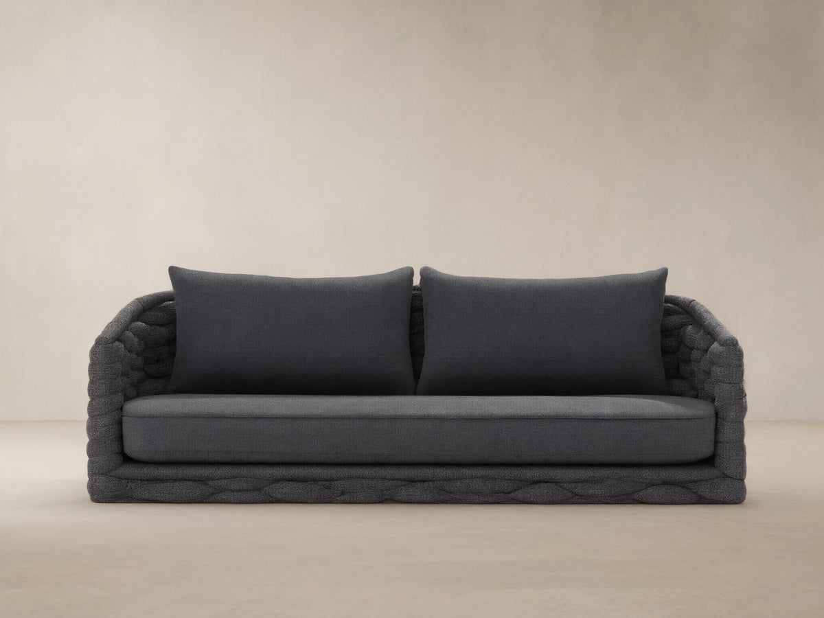 Melbourne Outdoor Sofa. Dark gray woven base, arms, & cushions. Against a light tan wall on a textured stone floor.