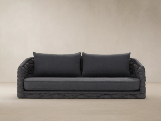 Melbourne Outdoor Sofa. Dark gray woven base, arms, & cushions. Against a light tan wall on a textured stone floor.