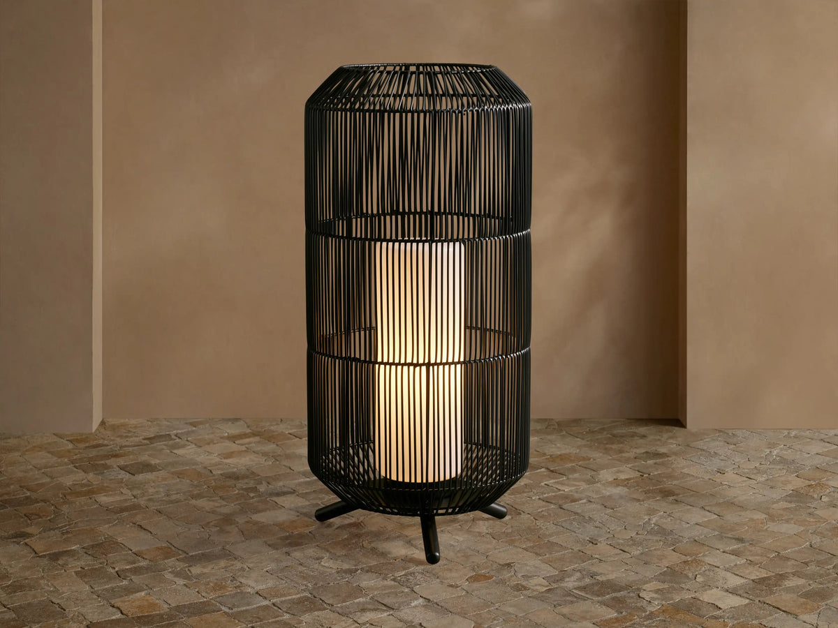 Black woven Mesa Outdoor Portable Lantern, tall cylinder, glowing light, on a textured stone floor against a tan wall.