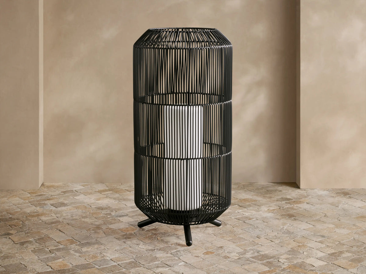 Black woven Mesa Outdoor Portable Lantern, tall cylinder, glowing light, on a textured stone floor against a tan wall.