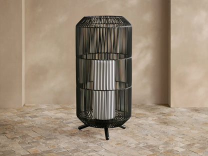 Black woven Mesa Outdoor Portable Lantern, tall cylinder, glowing light, on a textured stone floor against a tan wall.