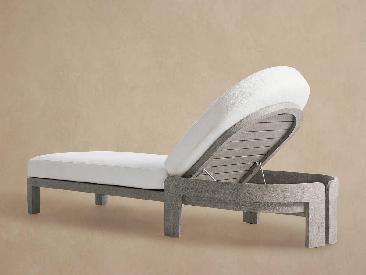 Milos Outdoor Chaise, grey weathered teak frame with white cushions. Side view, adjustable backrest raised, on a beige wall.