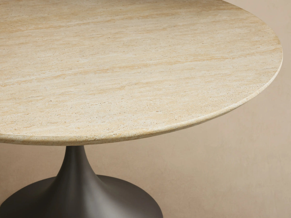 Outdoor round dining table with light beige textured stone top & dark gray flared pedestal Launette base on a tiled patio.