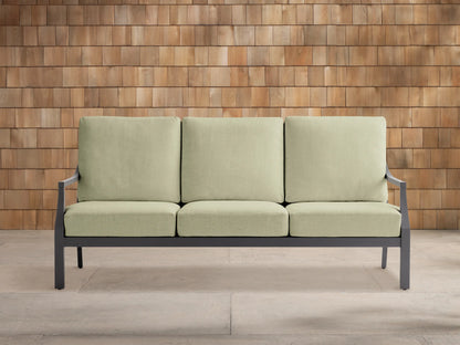 Montego outdoor aluminum sofa with dark gray frame and three light green Eden Sage cushions, on a light patio against a wood wall.