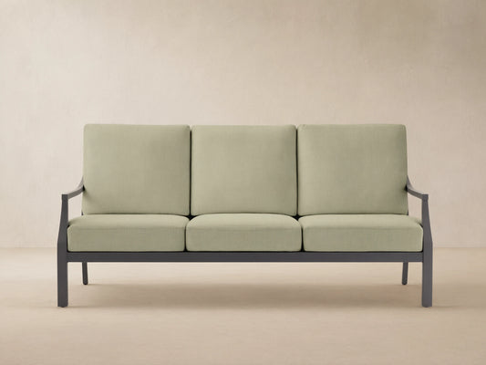 Montego outdoor aluminum sofa with dark gray frame and three light green Eden Sage cushions, on a light patio against a wood wall.