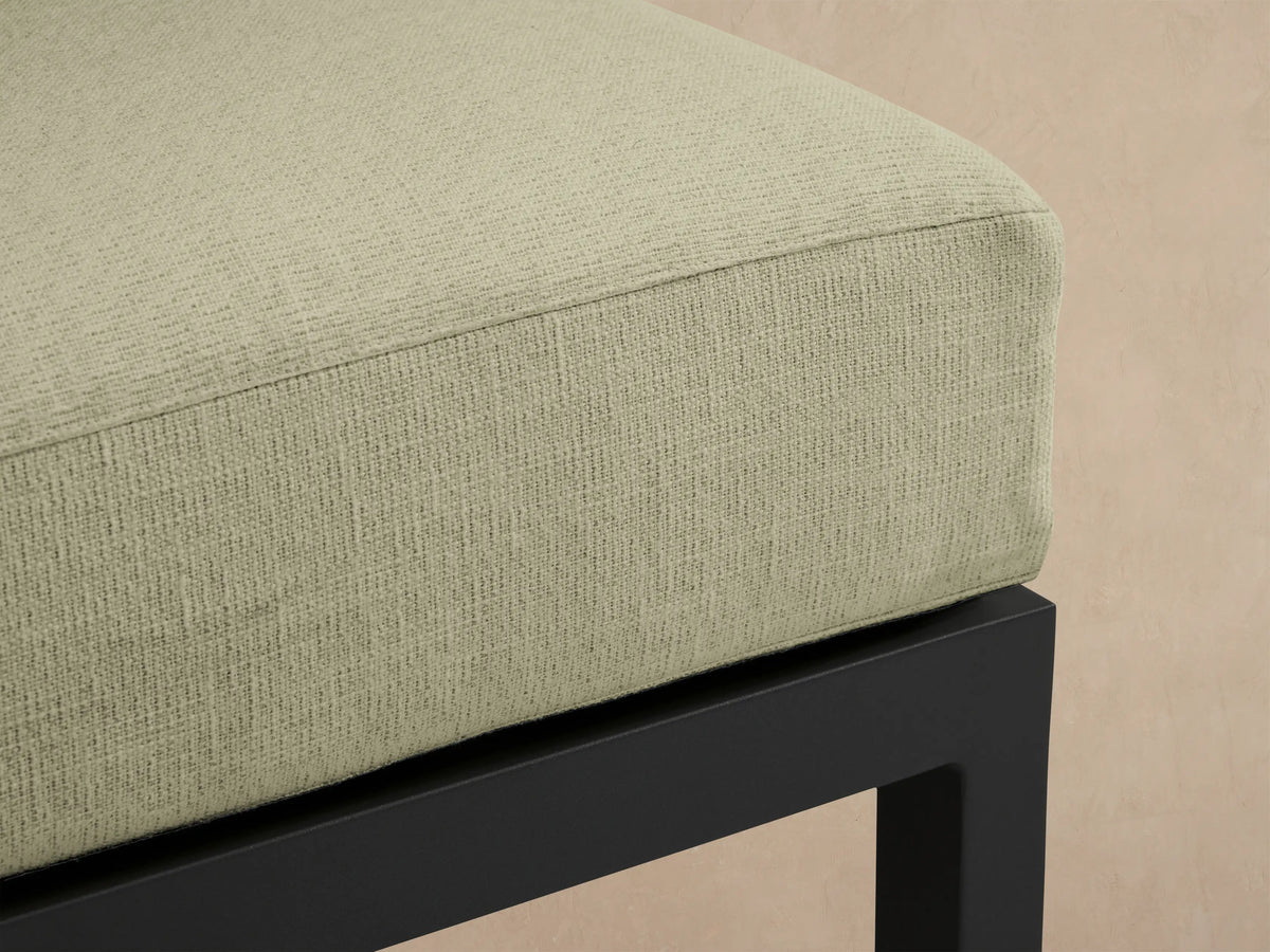 Montego Outdoor Aluminum Ottoman: square light green Eden Sage cushion on a dark gray frame, angled view against wood wall.