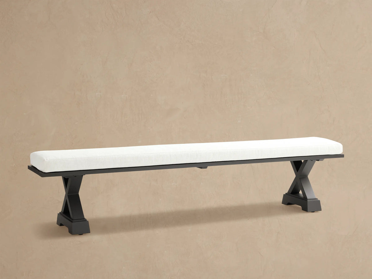Montego Outdoor Aluminum Dining Bench, front view. White cushion on black aluminum frame with angled trestle legs. Wooden wall background.