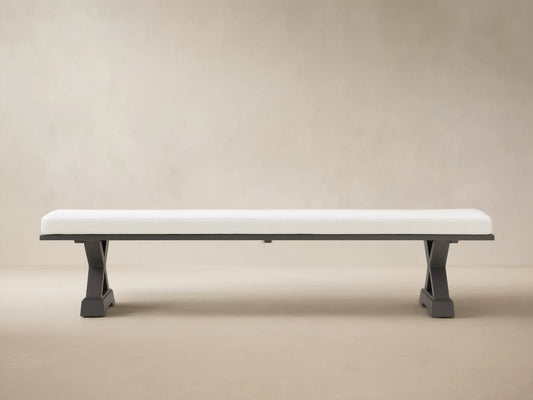 Montego Outdoor Aluminum Dining Bench, front view. White cushion on black aluminum frame with angled trestle legs. Wooden wall background.