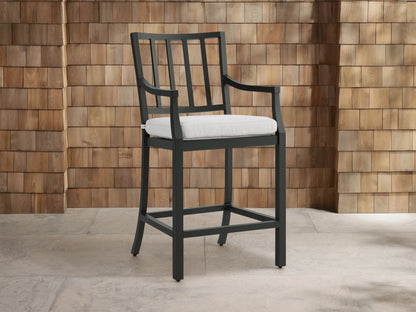 Montego Outdoor Aluminum Counter Stool, dark gray frame with slatted back and arms, white cushion. Angled on a patio.