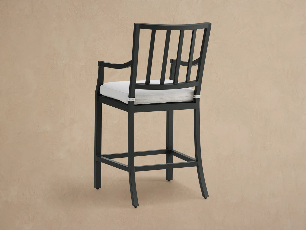 Close-up of Montego Outdoor Counter Stool. Dark aluminum frame with white seat cushion, visible armrest & slatted back.