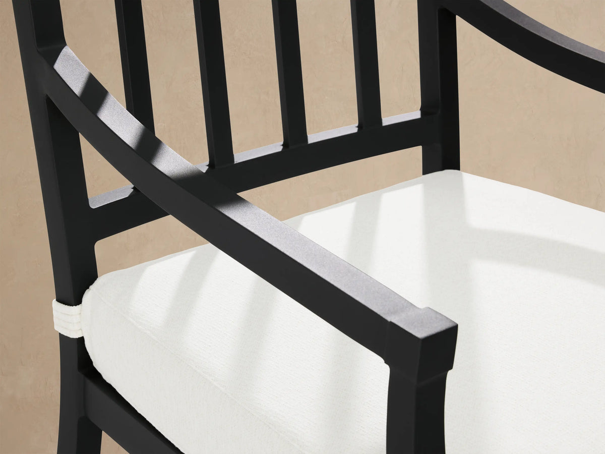 Montego Outdoor Aluminum Dining Arm Chair, black frame with white cushion, on light pavement against wood shingle wall.