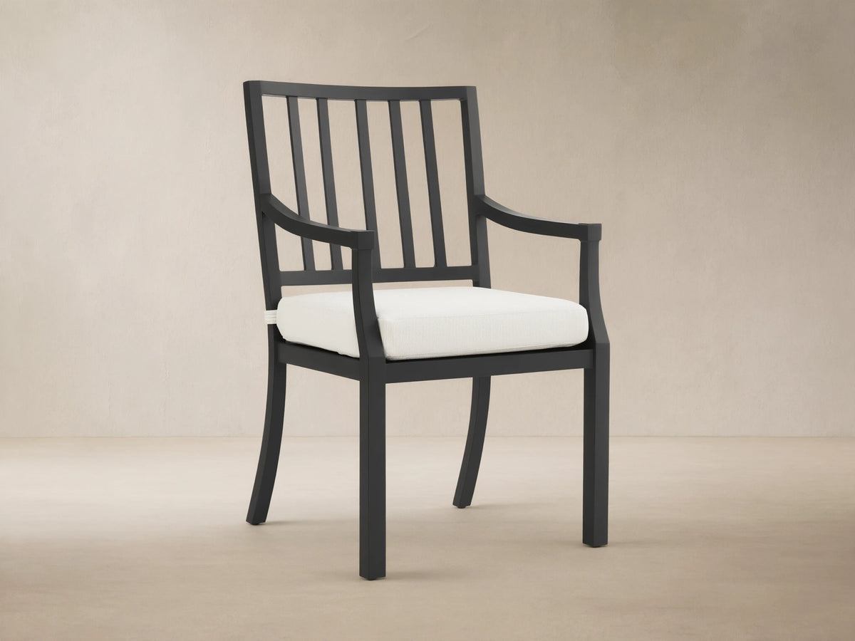 Montego Outdoor Aluminum Dining Arm Chair, black frame with slatted back, armrests & white cushion, shown angled on a patio.