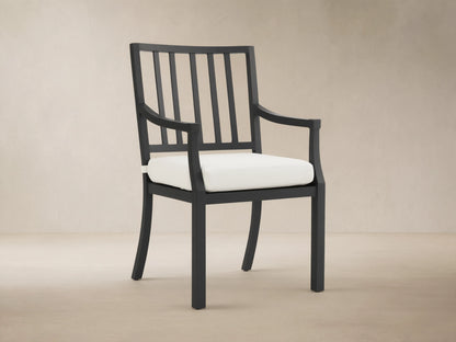 Montego Outdoor Aluminum Dining Arm Chair, black frame with slatted back, armrests & white cushion, shown angled on a patio.