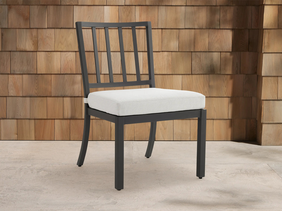 Montego Outdoor Aluminum Dining Side Chair with dark gray frame, slat back, and light cushion, on patio against wood wall.