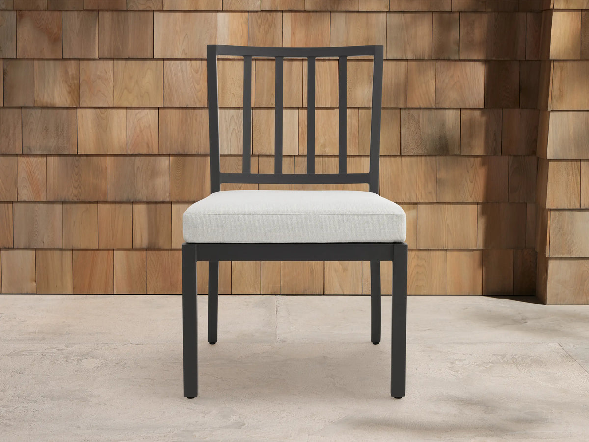 Montego Outdoor Aluminum Dining Side Chair with dark gray frame, slat back, and light cushion, on patio against wood wall.
