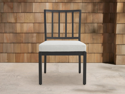 Montego Outdoor Aluminum Dining Side Chair with dark gray frame, slat back, and light cushion, on patio against wood wall.