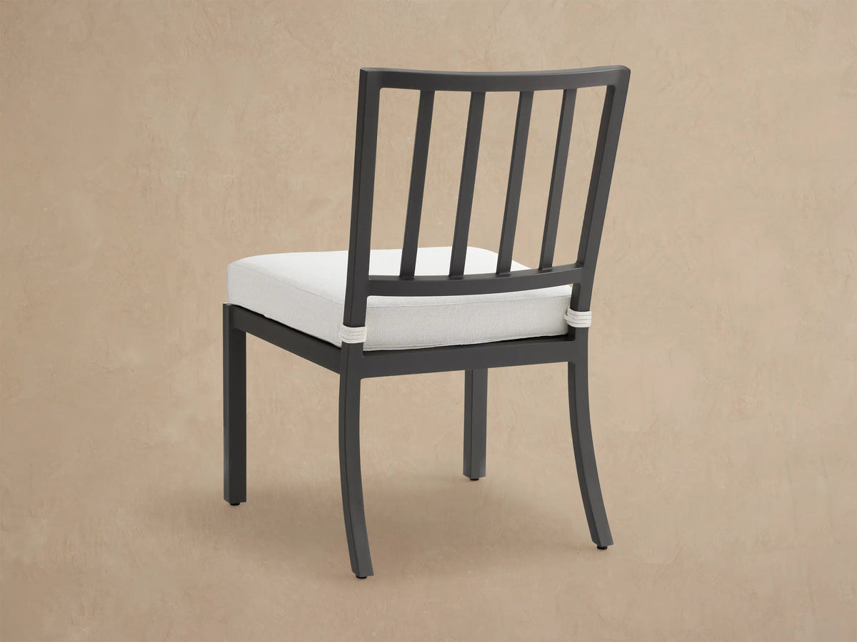 Angled close-up of a black aluminum Montego outdoor dining chair with a thick white cushion, showing rope tie and shadows.