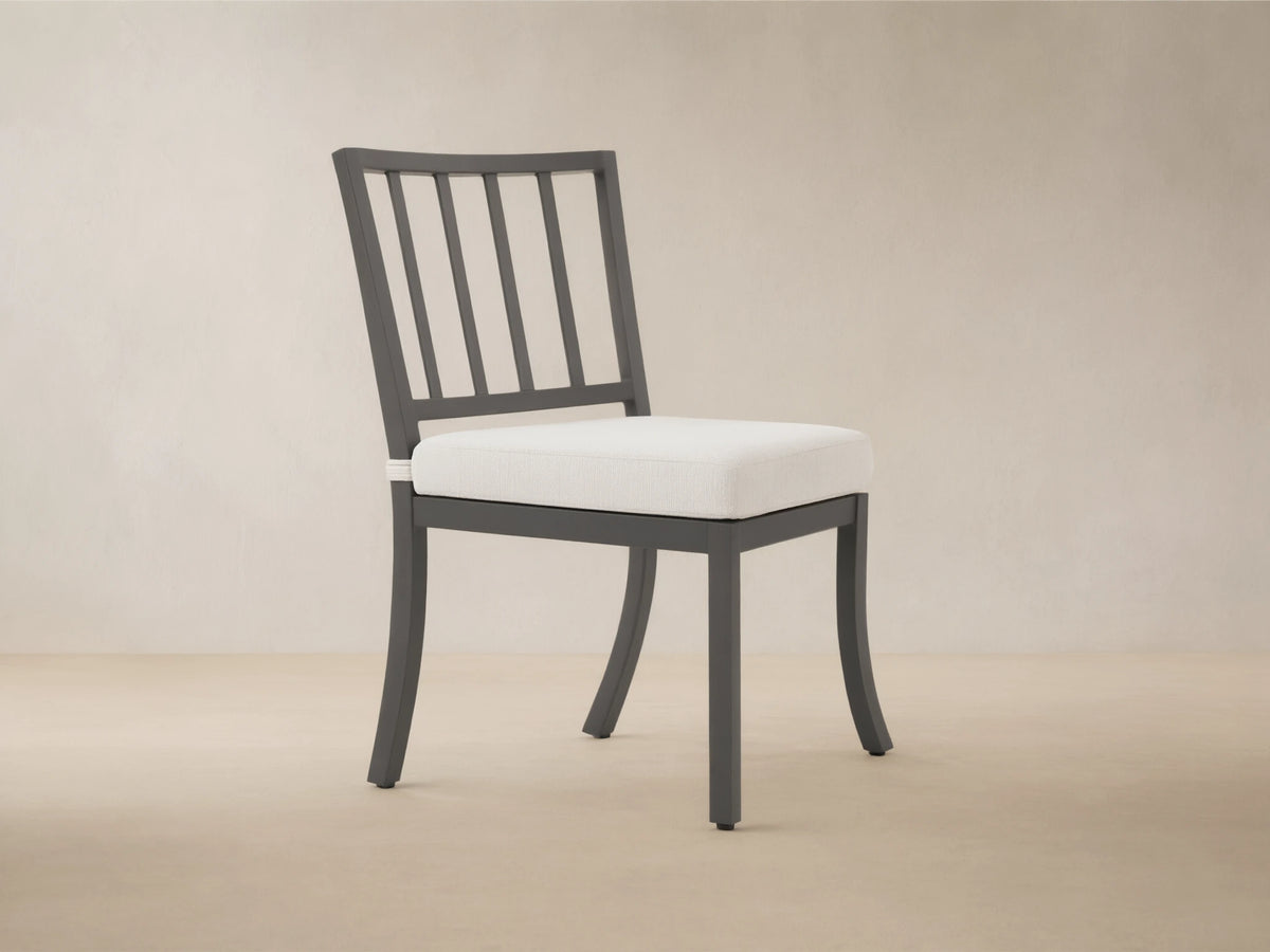 Montego Outdoor Aluminum Dining Side Chair with dark gray frame, slat back, and light cushion, on patio against wood wall.