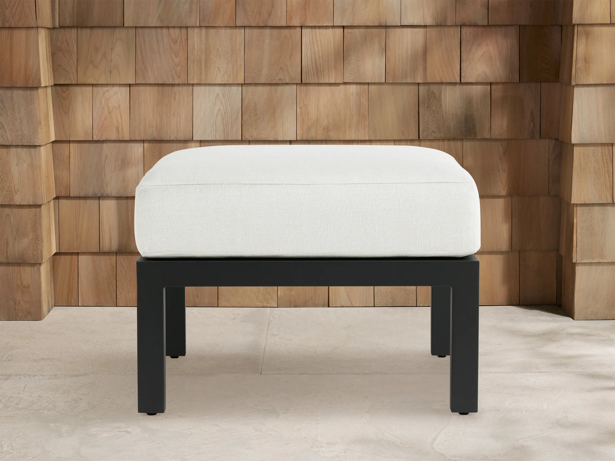 Montego Outdoor Aluminum Ottoman: white cushioned top on a dark gray rectangular aluminum base, in front of a cedar shingle wall.