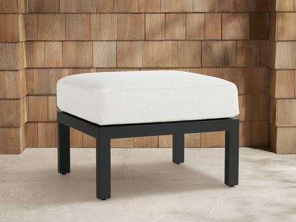 Montego Outdoor Aluminum Ottoman: white cushioned top on a dark gray rectangular aluminum base, in front of a cedar shingle wall.