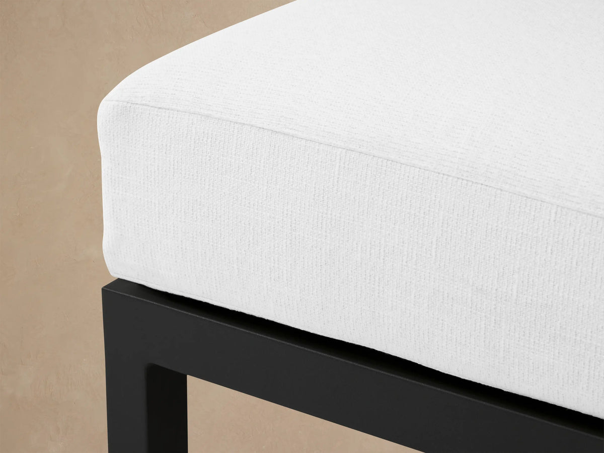 Montego Outdoor Aluminum Ottoman: square, dark gray frame with a thick off-white cushion, on light patio tiles against a wood shingle wall.