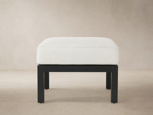 Montego Outdoor Aluminum Ottoman: white cushioned top on a dark gray rectangular aluminum base, in front of a cedar shingle wall.