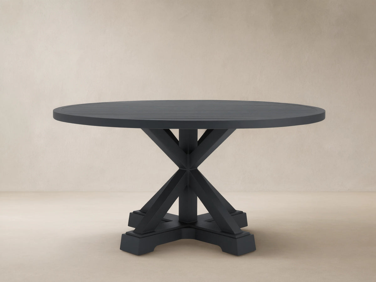 Montego Outdoor Aluminum Round Dining Table, charcoal gray, with a slatted top, X-pedestal base, on patio, wood shingle wall.