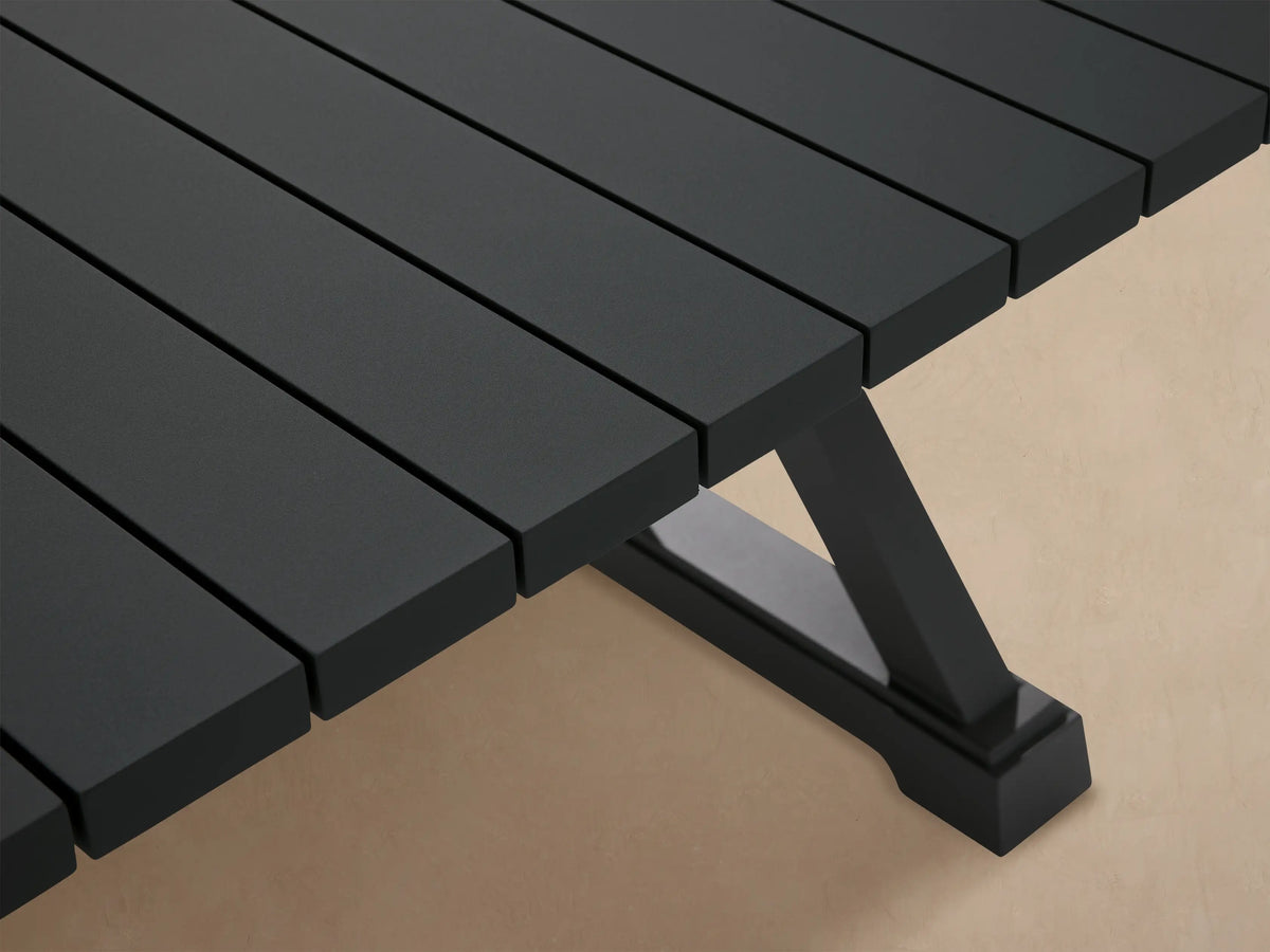 Dark gray Montego Outdoor Aluminum Dining Table with a slatted top and X-shaped legs on a patio with wood shingles.