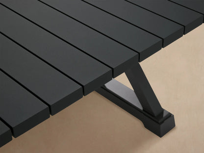 Dark gray Montego Outdoor Aluminum Dining Table with a slatted top and X-shaped legs on a patio with wood shingles.