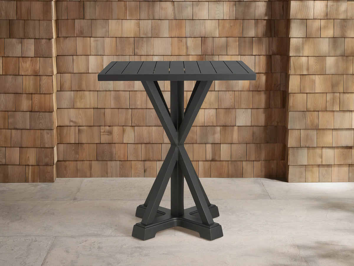 Dark gray Montego Outdoor Aluminum Bar Table with a slatted square top and an X-shaped base, on concrete before a shingle wall.