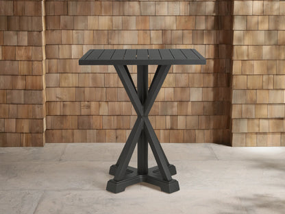 Dark gray Montego Outdoor Aluminum Bar Table with a slatted square top and an X-shaped base, on concrete before a shingle wall.
