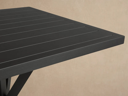 Dark gray Montego Outdoor Aluminum Bar Table with a slatted square top and an X-shaped base, on concrete before a shingle wall.