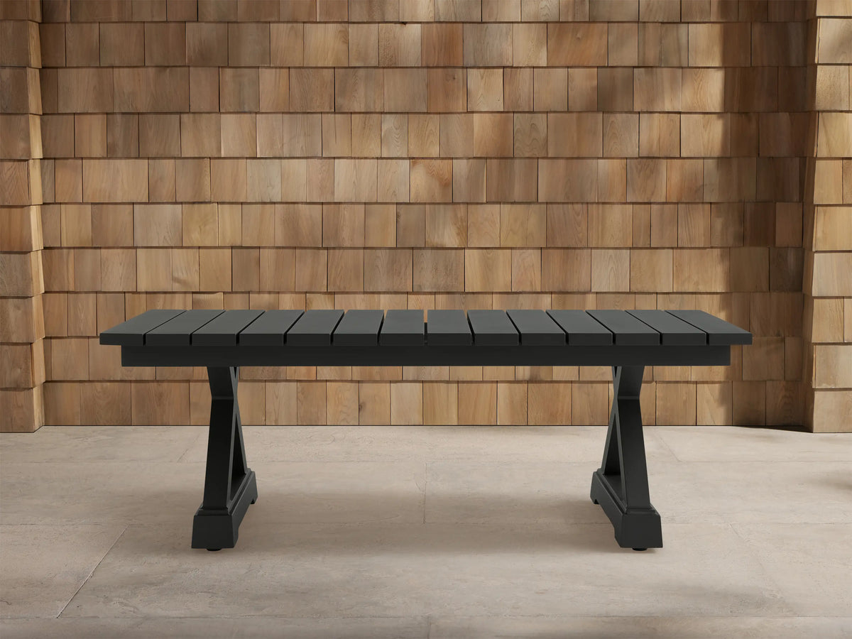 Dark gray Montego Outdoor Aluminum Coffee Table with slatted top, trestle base, on light patio tiles against wood shingle wall.