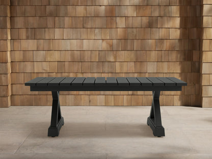 Dark gray Montego Outdoor Aluminum Coffee Table with slatted top, trestle base, on light patio tiles against wood shingle wall.