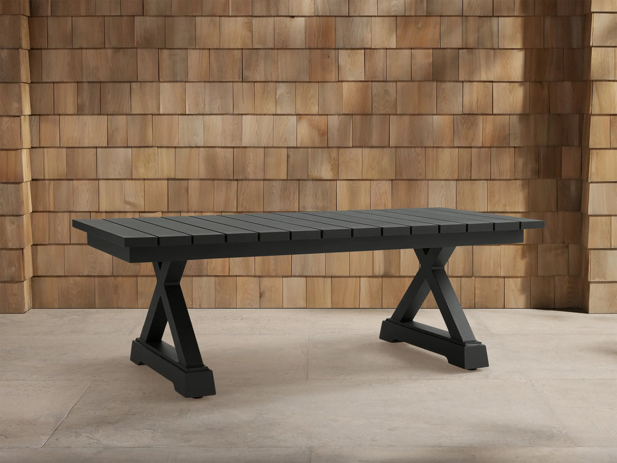 Dark gray Montego Outdoor Aluminum Coffee Table with slatted top, trestle base, on light patio tiles against wood shingle wall.