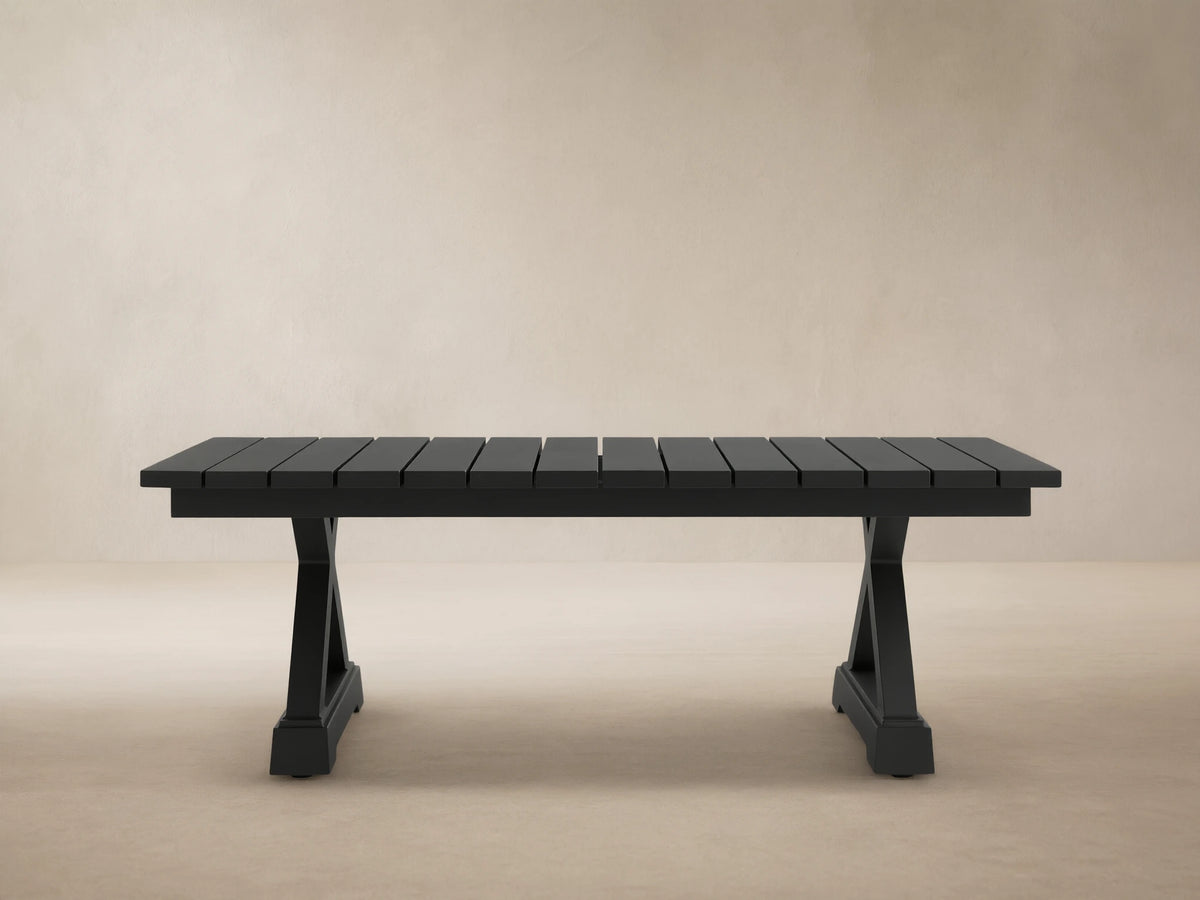 Dark gray Montego Outdoor Aluminum Coffee Table with slatted top, trestle base, on light patio tiles against wood shingle wall.