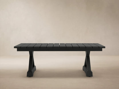 Dark gray Montego Outdoor Aluminum Coffee Table with slatted top, trestle base, on light patio tiles against wood shingle wall.