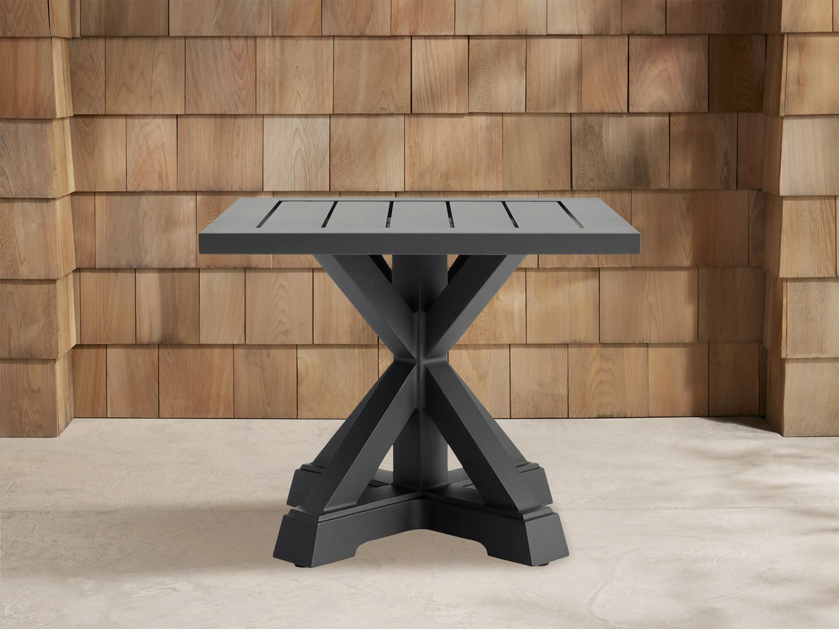 Dark grey Montego Outdoor Aluminum End Table with slatted square top, cross-shaped base, against a wood shingle wall.