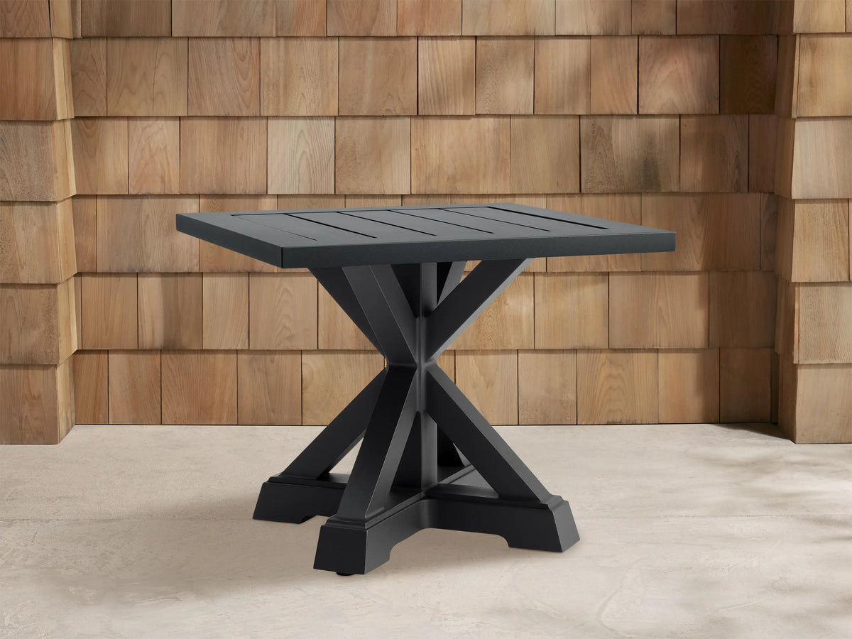 Dark grey Montego Outdoor Aluminum End Table with slatted square top, cross-shaped base, against a wood shingle wall.