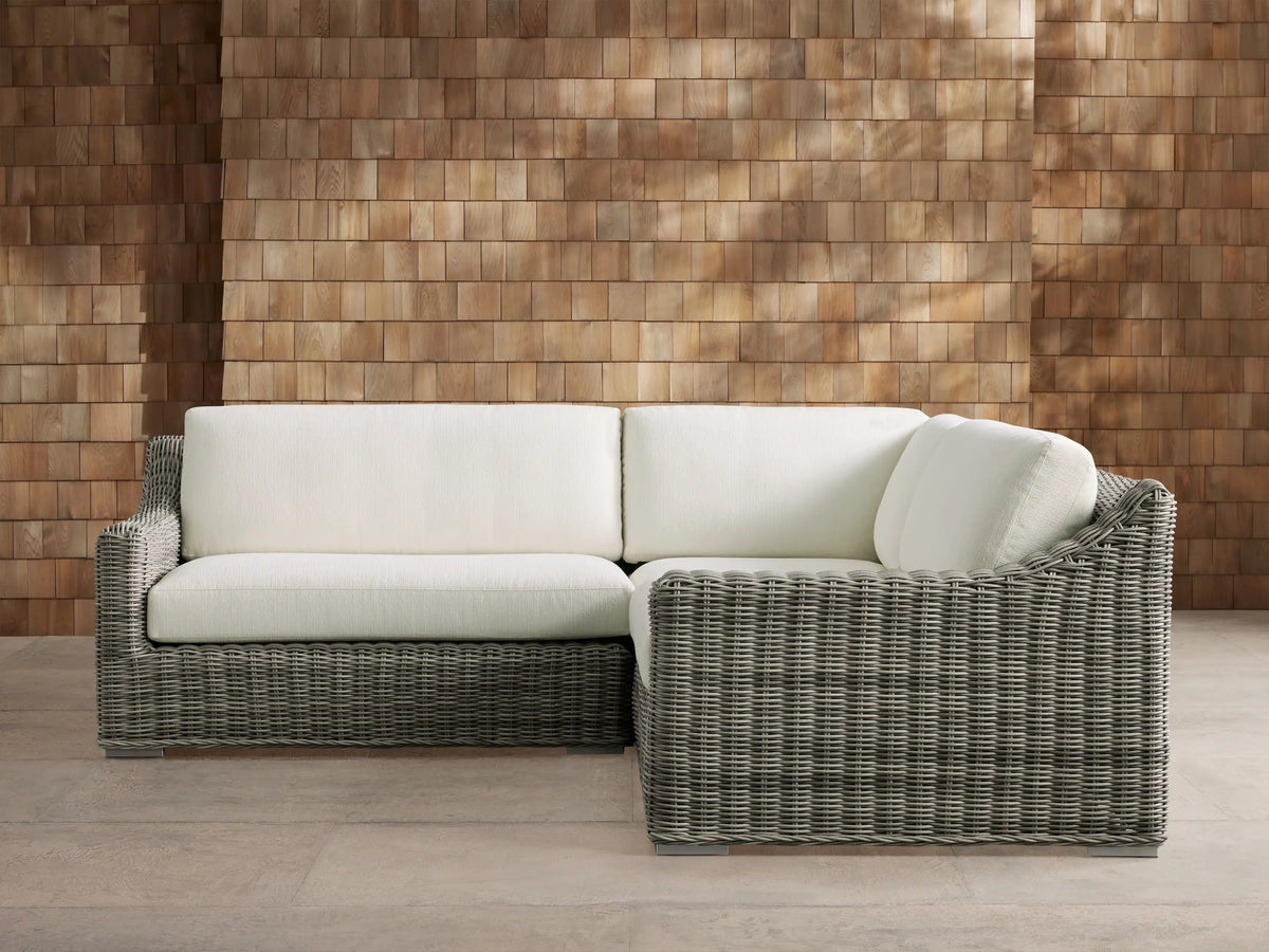 Montauk Outdoor Three-Piece Sectional, L-shaped, gray handwoven resin wicker with white cushions, on patio, against wood wall.