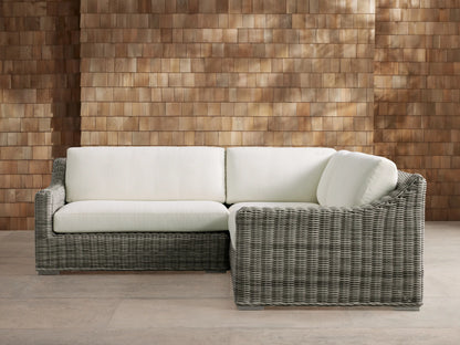 Montauk Outdoor Three-Piece Sectional, L-shaped, gray handwoven resin wicker with white cushions, on patio, against wood wall.