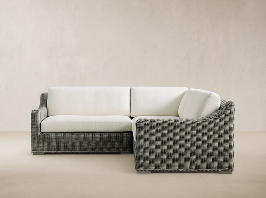 Montauk Outdoor Three-Piece Sectional, L-shaped, gray handwoven resin wicker with white cushions, on patio, against wood wall.