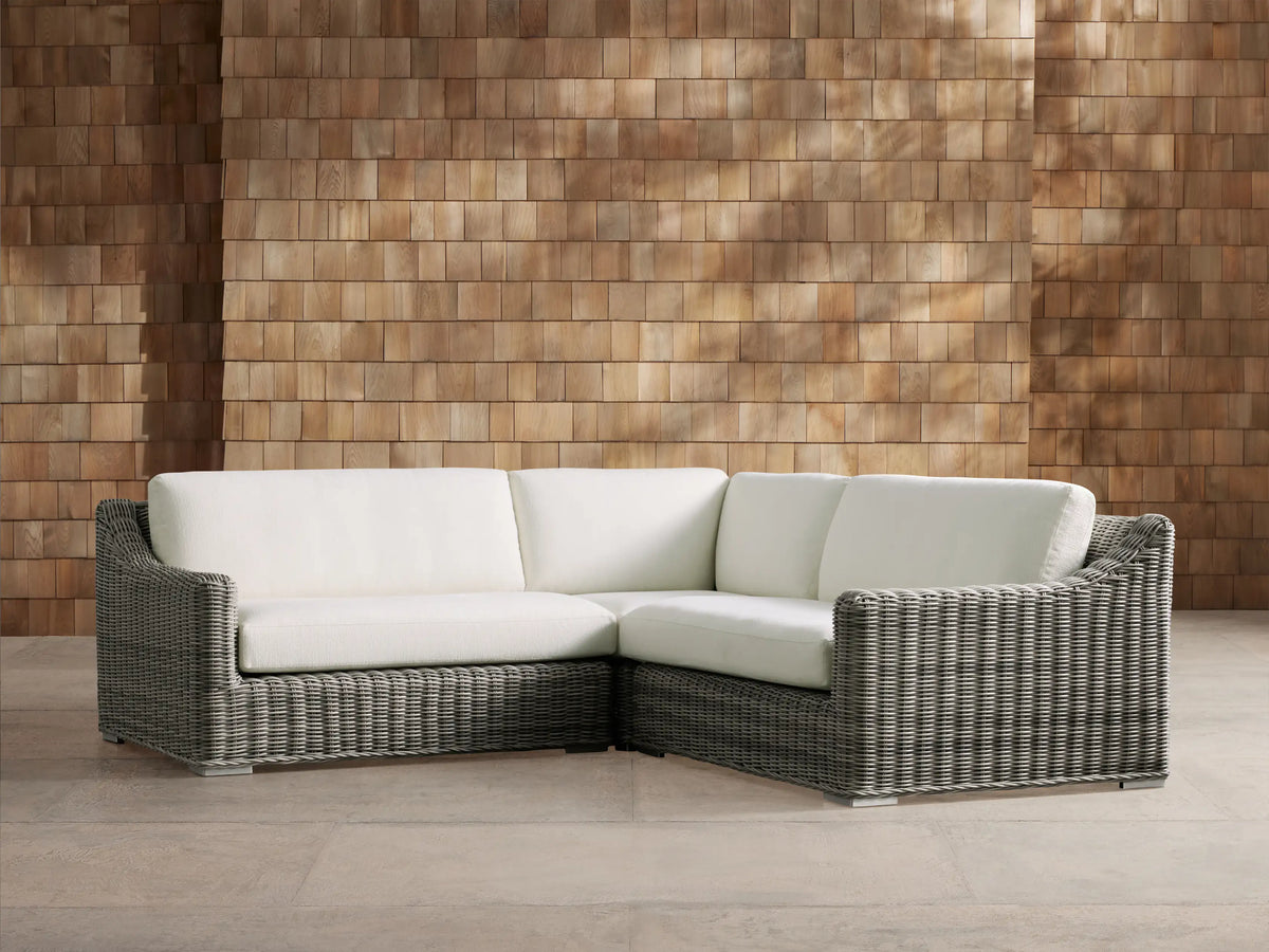 Montauk Outdoor Three-Piece Sectional, L-shaped, gray handwoven resin wicker with white cushions, on patio, against wood wall.