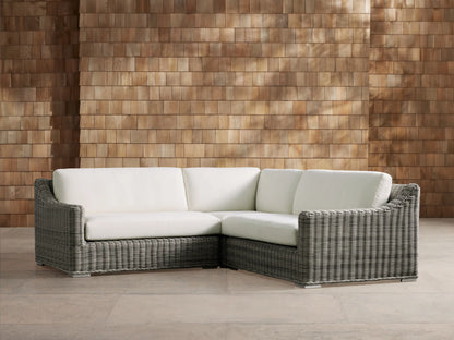 Montauk Outdoor Three-Piece Sectional, L-shaped, gray handwoven resin wicker with white cushions, on patio, against wood wall.