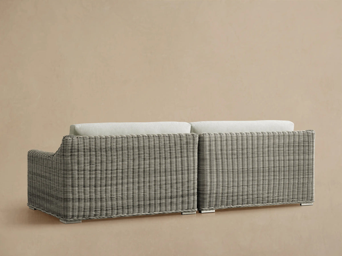 Rear view of the light gray woven wicker Montauk Outdoor Two-Piece Sectional, with off-white cushions and stainless-steel feet.