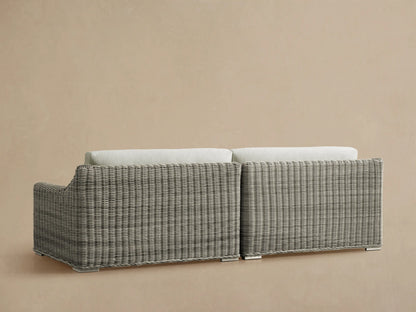 Rear view of the light gray woven wicker Montauk Outdoor Two-Piece Sectional, with off-white cushions and stainless-steel feet.