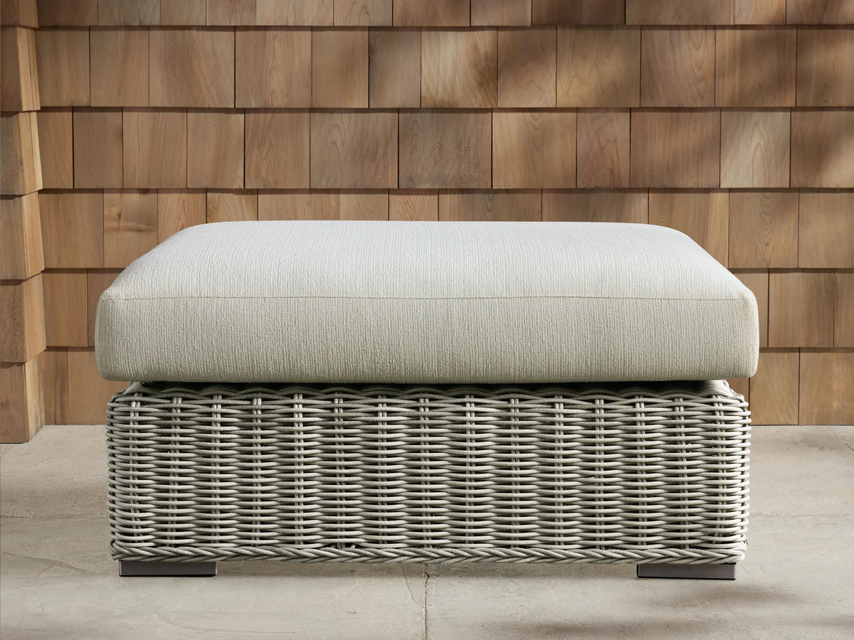 Montauk Outdoor Ottoman: pale sage woven resin wicker base, light textured cushion. On patio, wood shingle background.