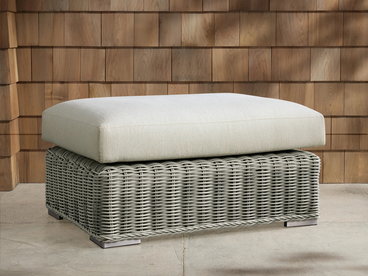 Montauk Outdoor Ottoman: pale sage woven resin wicker base, light textured cushion. On patio, wood shingle background.