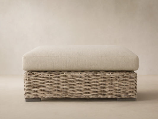 Montauk Outdoor Ottoman: pale sage woven resin wicker base, light textured cushion. On patio, wood shingle background.